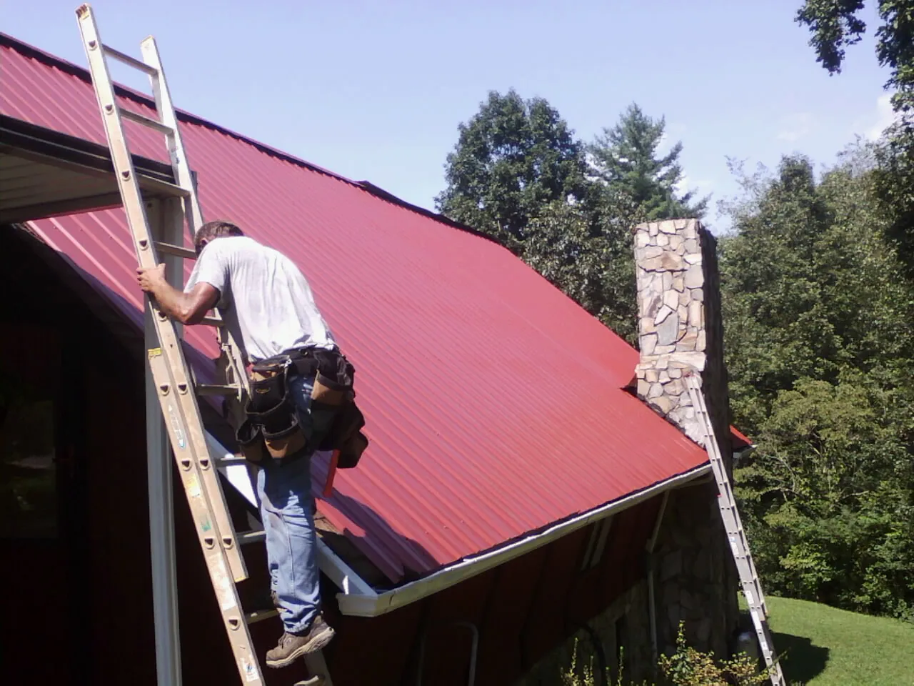 Roofer on a ladder beside a red metal roof and stone chimney — Soffit & Fascia Repair by Roofing Brothers in Spanish Fork